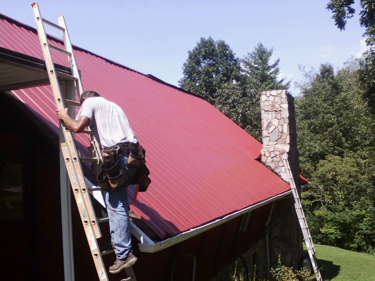 Roofer on a ladder beside a red metal roof and stone chimney — Soffit & Fascia Repair by Roofing Brothers in Suttons Bay